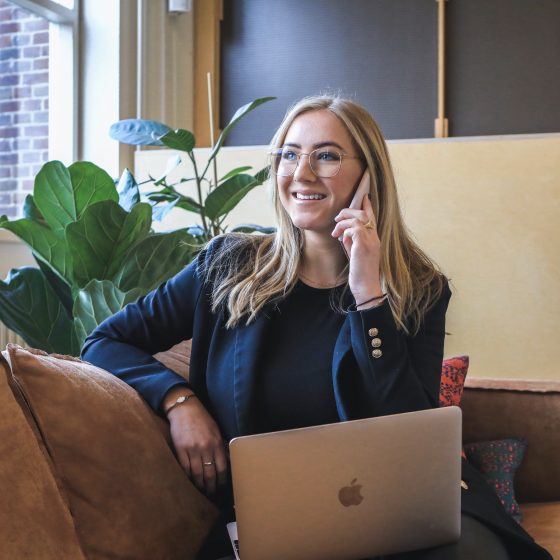 A woman is talking to someone with her phone and holding a laptop on her knee
